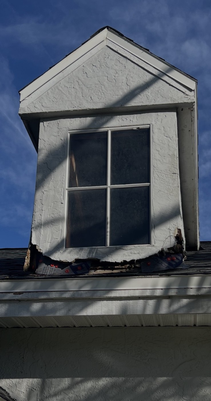 A roof dormer with rotted trim and crumbling stucco along the bottom